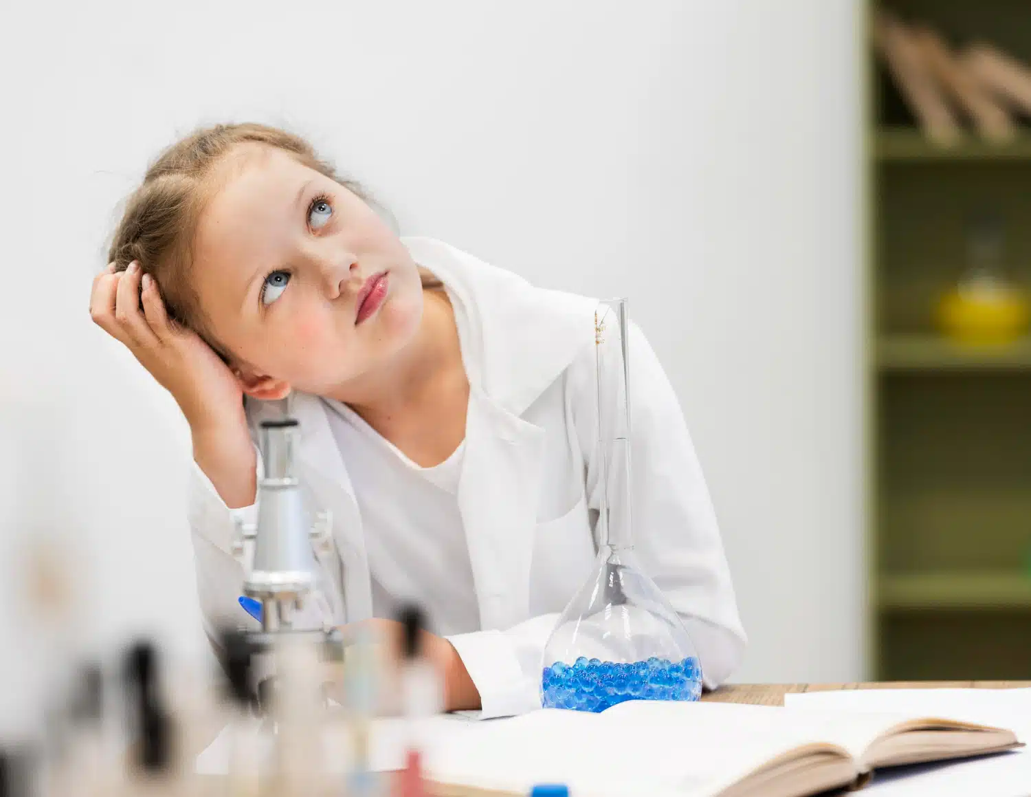 A young girl in a white lab coat sits at a desk with science equipment and an open book, resting her head on her hand and looking up thoughtfully.