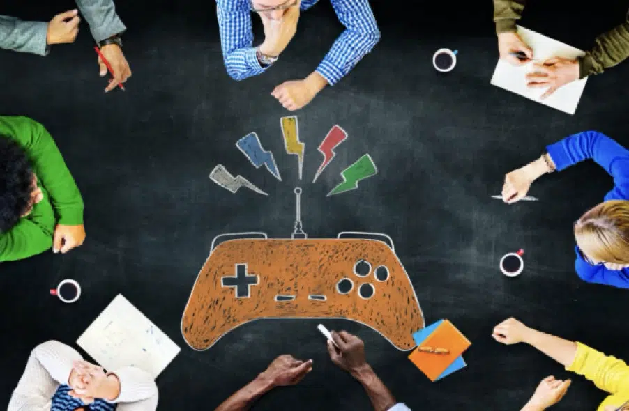 People sitting around a table with a large drawing of a video game controller on a blackboard surface, holding notebooks, pens, and coffee cups.
