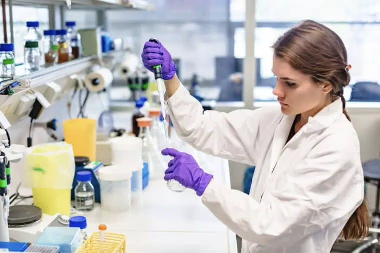 A woman in a lab coat and purple gloves uses a pipette to transfer liquid in a laboratory setting with equipment and bottles on the bench.
