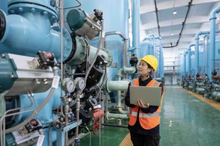 An engineer wearing a hard hat and safety vest inspects industrial machinery, holding a laptop in a large facility with blue pipes and equipment.