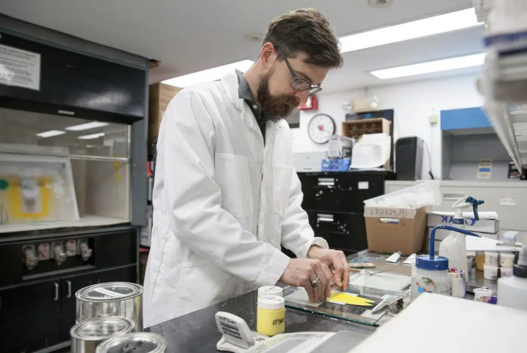 A man in a white lab coat works with samples on a glass tray in a laboratory filled with equipment and supplies.