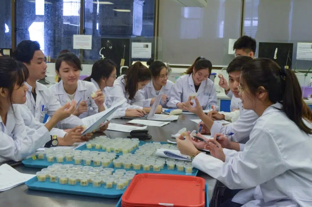 A group of students in lab coats sit around a table in a laboratory, examining samples and taking notes, with trays of specimen containers in front of them.