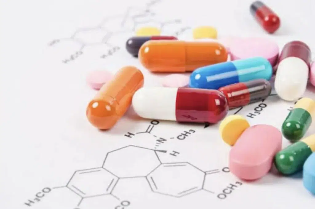 Assorted pills and capsules are scattered on a sheet of paper with printed chemical structures.