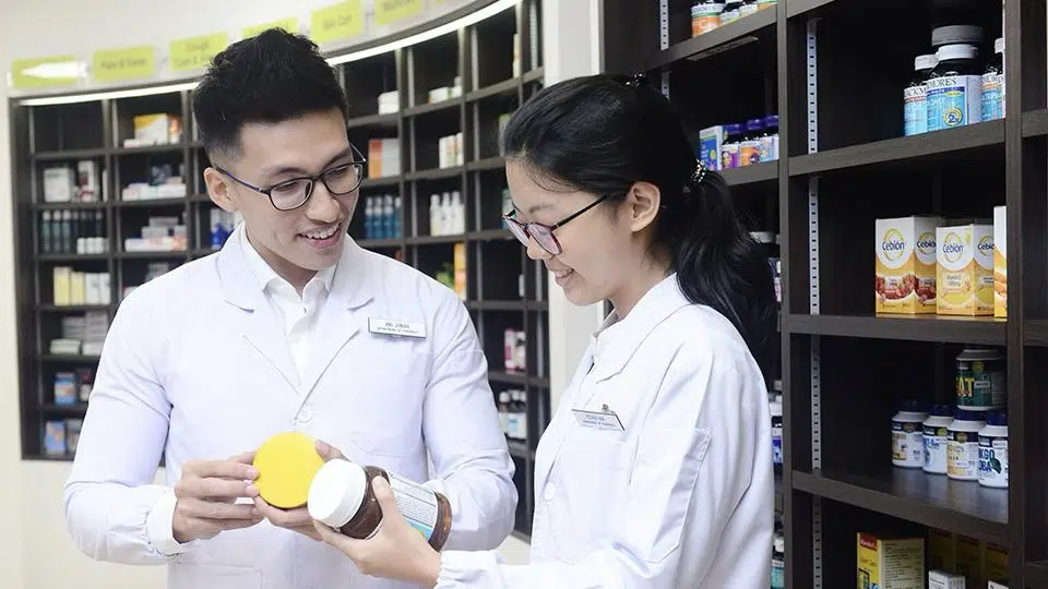 Two pharmacists in white coats discuss a product while standing in a pharmacy, surrounded by shelves stocked with various medicines and supplements.