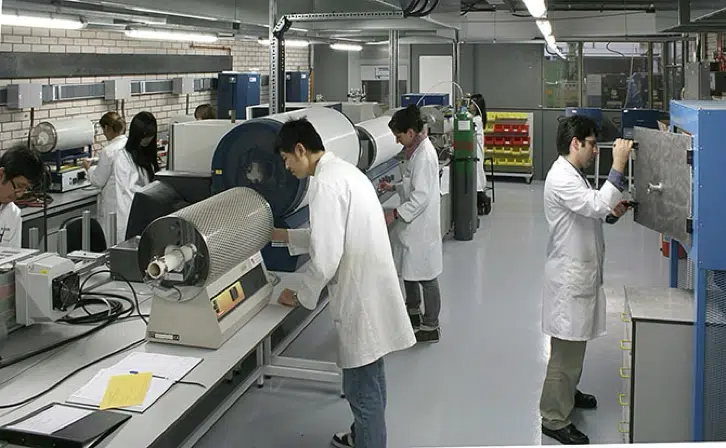 Several people in lab coats work with scientific equipment in a laboratory setting with various machines, tools, and storage units visible.