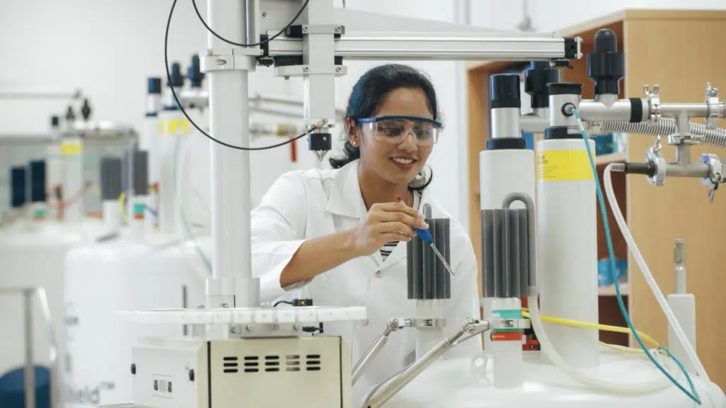 A scientist wearing safety goggles and a lab coat adjusts equipment in a laboratory filled with large scientific instruments and machinery.