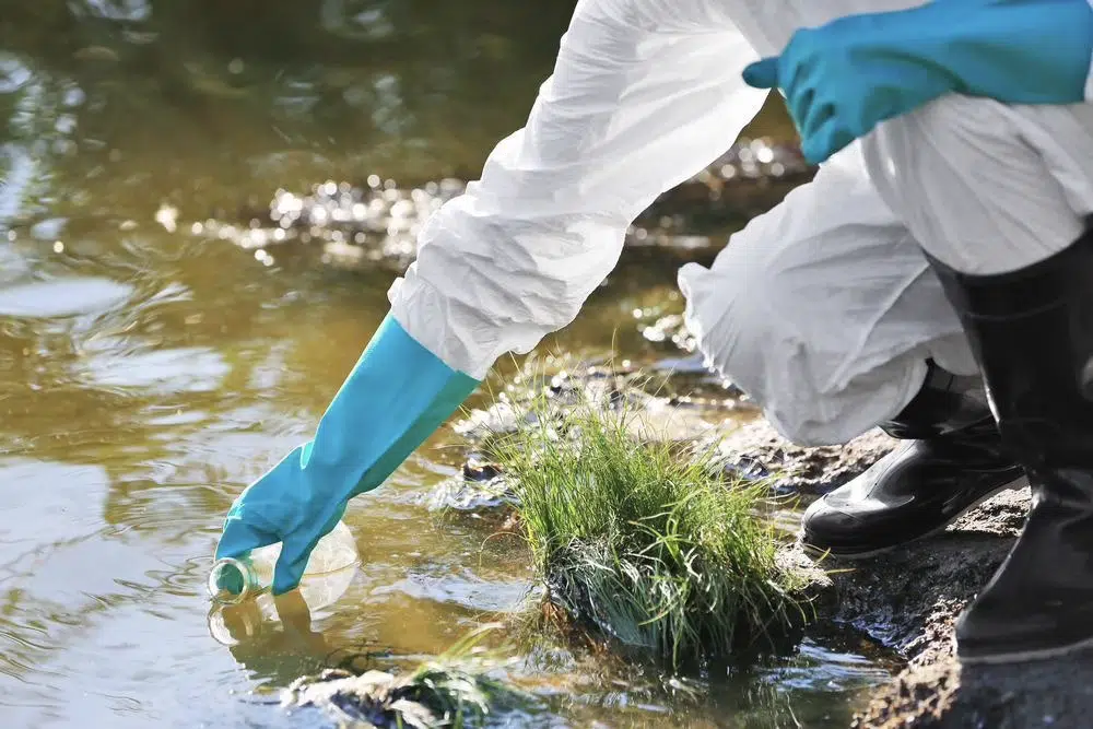 Person in protective gear and blue gloves collects a water sample from a riverbank using a clear bottle.