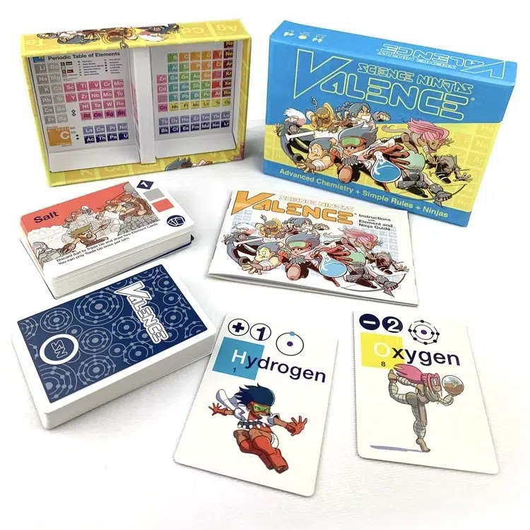 The image shows the card game "Science Ninjas: Valence," including the box, rulebook, a reference card, and several playing cards featuring elements like Hydrogen, Oxygen, and Salt.