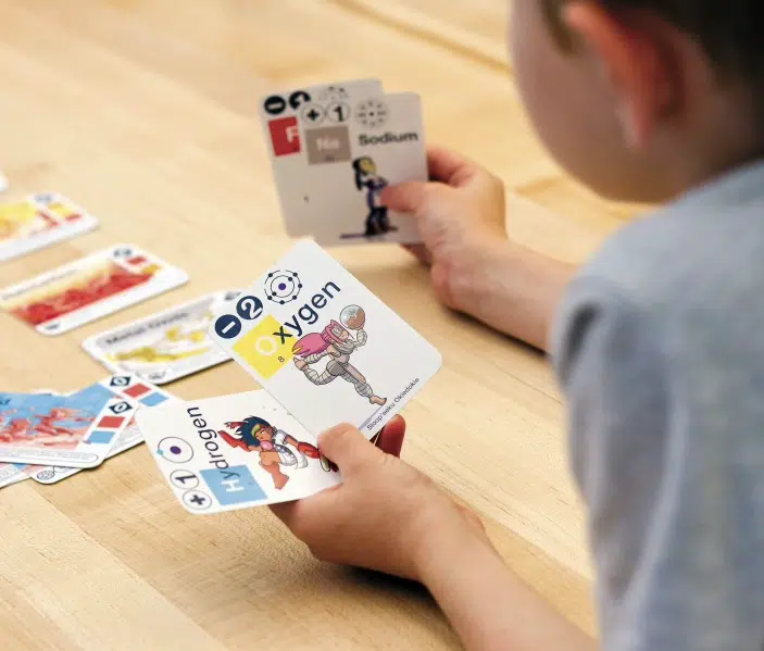 A child sits at a wooden table holding cards labeled "Hydrogen" and "Oxygen" while playing a science-themed card game.