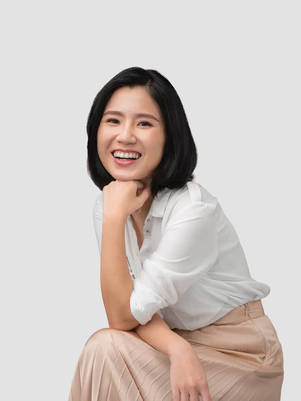 A woman with short black hair wearing a white blouse and beige skirt sits and smiles at the camera against a plain light background, presenting a friendly face for science tuition.