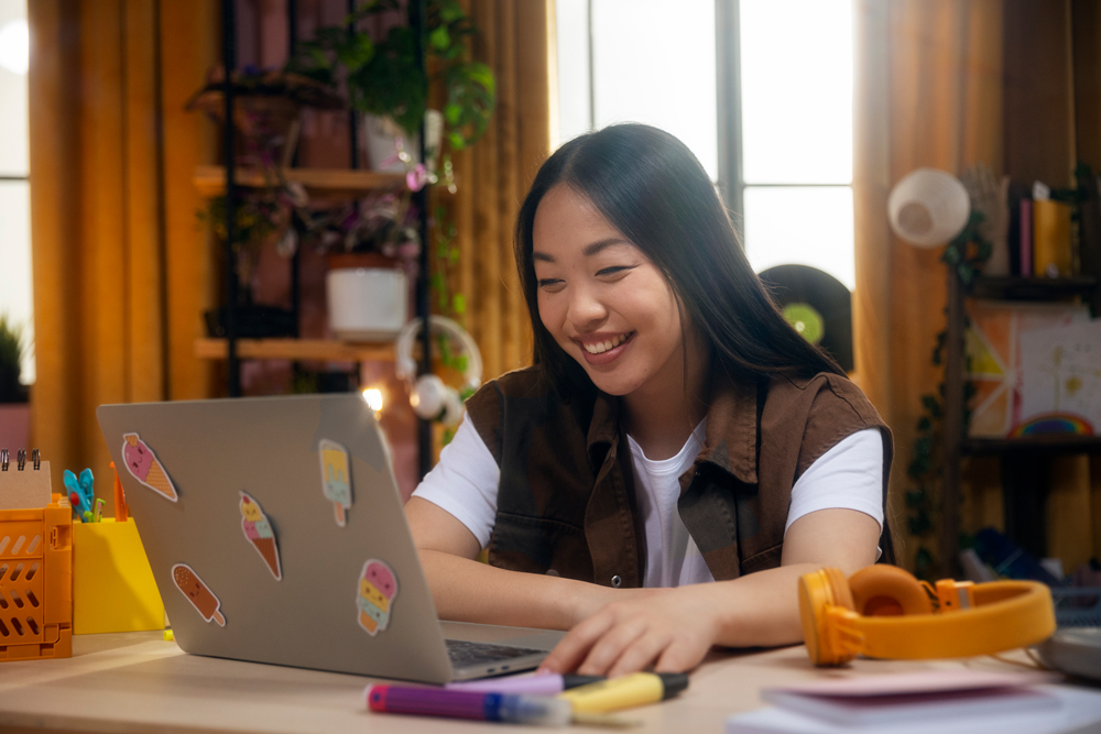A young woman sits at a desk, smiling at her laptop decorated with stickers, surrounded by yellow headphones, pens, and a notebook as she enjoys Science Tuition in a brightly lit room.