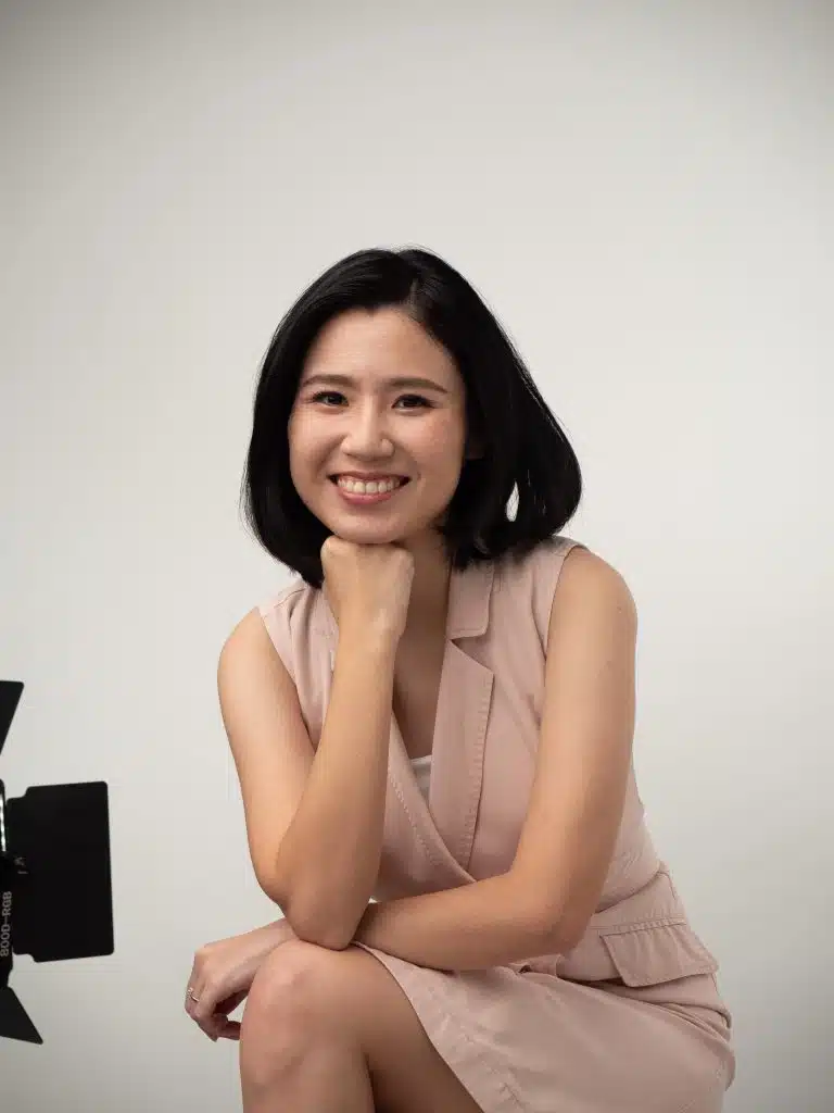 A woman with short black hair wearing a sleeveless beige dress sits on a stool, smiling at the camera with her chin resting on her hand. A studio light is partially visible.
