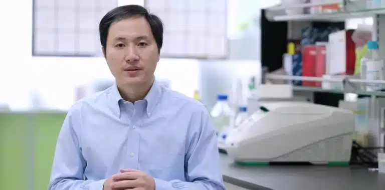 A man in a light blue shirt stands in a laboratory, with scientific equipment, supplies, and a periodic table visible on shelves and a counter behind him.