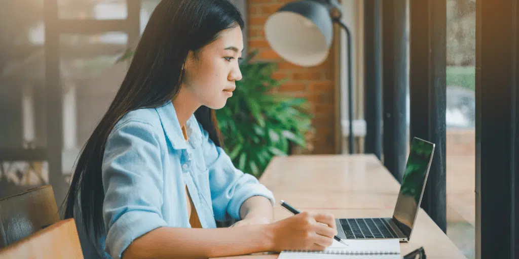 A young woman sits at a desk by a window, taking notes in a notebook while looking at a laptop.