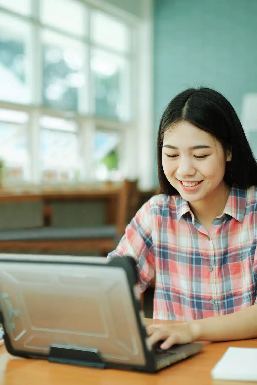 A young woman in a plaid shirt sits at a table, smiling and working on a laptop in a bright room with large windows.