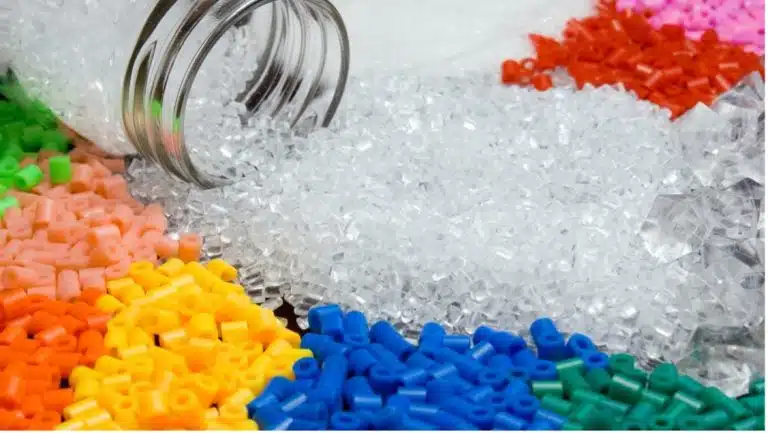 A glass jar spills clear plastic pellets onto a surface surrounded by separate piles of colored plastic pellets in red, orange, yellow, green, and blue—each representing different functional groups.