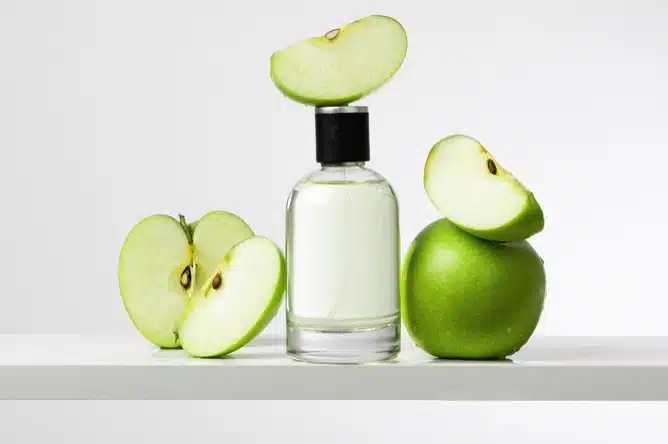 A clear perfume bottle is placed between a whole green apple and green apple slices on a white surface, with an apple slice balanced on top of the bottle, highlighting the freshness inspired by the functional groups in fruity fragrances.