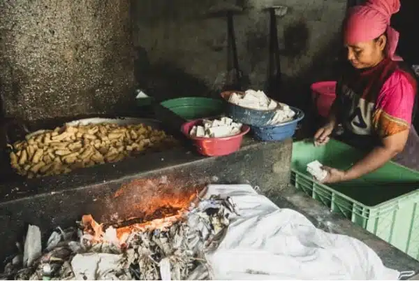 A woman wearing a pink headscarf prepares food beside a wood-fired stove, where the polymerisation of ingredients creates delicious cooked items arranged on trays beside her.
