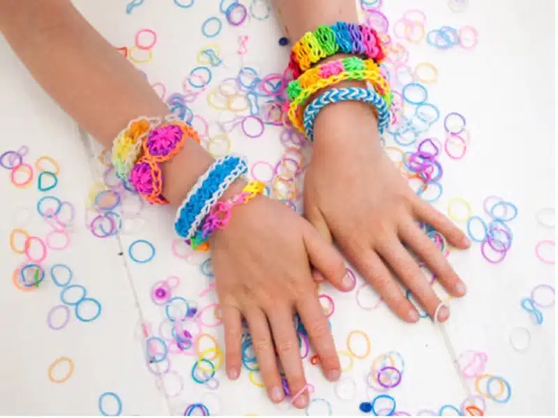 Two hands wearing multiple colorful loom band bracelets, created through polymerisation, rest on a white surface scattered with loose rubber bands.
