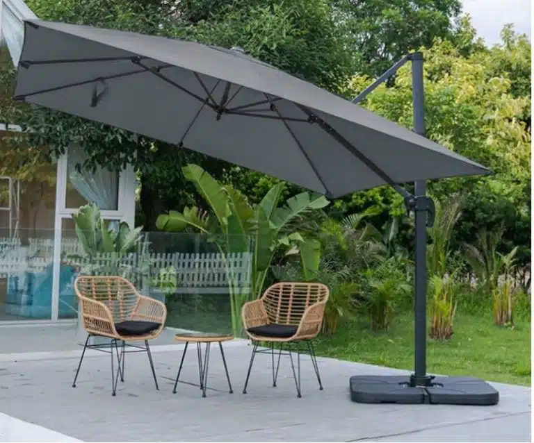 Two wicker chairs and a small round table sit beneath a large gray cantilever umbrella on the patio, featuring lush greenery and a building in the background—a serene spot to unwind after studying polymerisation.